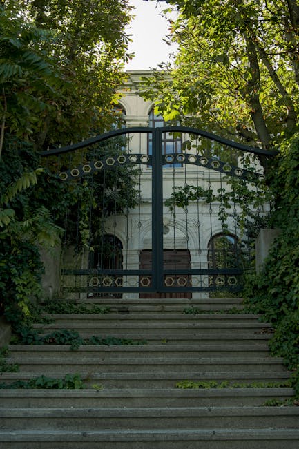 A set of concrete steps leading up to a decorative metal gate with circular and linear patterns, flanked by lush green foliage from trees and bushes on both sides. The gate opens to a driveway or courtyard area in front of a white building with arched windows, partially obscured by trees. The scene is illuminated by natural daylight, with shadows cast by the leaves. This outdoor setting suggests a residential property with a gated entrance, and the image captures the front approach during a bright day. Man and Van Forest Gate are specialists in home relocation, including furniture transport and packing, often involving gates and entrance accessibility as part of the moving process.