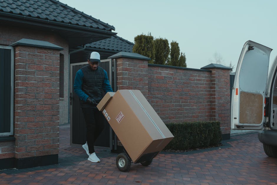 A man wearing a blue and black jacket, black pants, white sneakers, and a white cap is loading a large cardboard box onto a wheeled trolley outside a residential property during an eventual home relocation process. The box, secured with packing tape, rests on the trolley, which is positioned on a brick-paved driveway. He is standing near a brick wall with a dark grey gate, on the left side of the image, which provides access to the property. To the right, a white van with its rear door open is parked on the driveway, ready for furniture transport and packing and moving activities associated with professional house removals. In the background, there are tall, green bushes and a house with a dark tiled roof and window shutters. The scene is lit with natural daylight, and the environment suggests an organized process of loading household items for a residential move, typical of services offered by Man and Van Forest Gate.