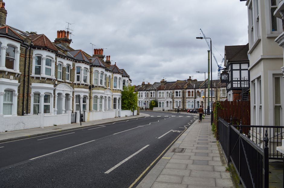A quiet residential street in a Victorian-style neighbourhood with row houses featuring bay windows, decorative brickwork, and chimney stacks. The street is lined with paved sidewalks, some with metal railings, and has a few trees along the pavement. Overcast grey skies create diffuse lighting, with no visible vehicles or pedestrians. The scene is captured from sidewalk level, showing the front facades of houses on both sides of the road, and extending into the distance where a crane is visible, indicating nearby construction work. The environment suggests a typical setting for house removals or furniture transport, supported by the calm, spacious street layout and residential architecture, aligning with the services offered by Man and Van Forest Gate, as described in the bulky item removals guide for Forest Gate Wanstead Flats.