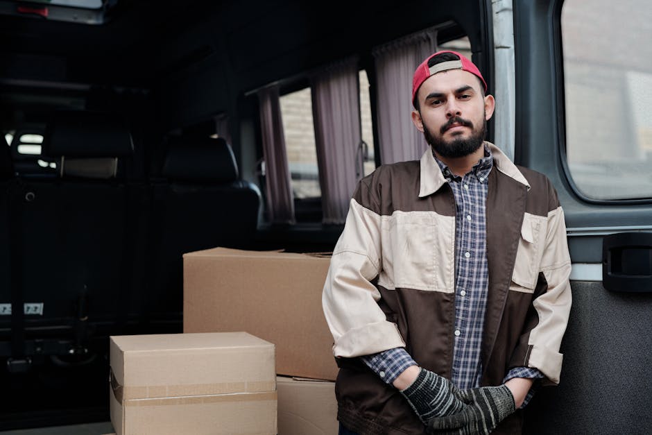Two men sitting at the rear end of a van with its back doors open, surrounded by packed cardboard boxes and wrapped furniture, possibly large appliances or household goods, ready for a home relocation