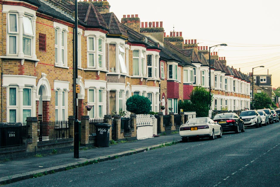 A row of traditional terraced houses in Forest Gate with brick facades, white window frames, and small front gardens enclosed by low brick walls with metal railings. Some properties have bay windows and decorative architectural details. Parked cars, including a white vehicle closest to the camera, line the street pavement. A black wheelie bin is situated near the curb, and a street lamp extends over the road. The scene appears to be taken during daylight with soft, natural lighting. This image exemplifies typical residential housing in Forest Gate, capturing details relevant to home relocation and moving logistics, where the presence of parked vehicles and street layout are important for planning removals services. Man and Van Forest Gate conducts furniture transport and packing tasks in this area, supporting efficient house removals and adherence to local permit rules for Forest Gate removals, as discussed on the company's webpage about Newham council permit requirements.