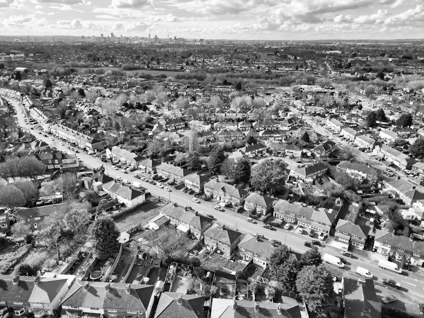 An aerial black-and-white photograph showing a residential neighbourhood in Forest Gate, East London, with a variety of terraced houses, semi-detached homes, and detached properties. The image captures a main street lined with parked cars and driveways, with trees and gardens visible throughout the area. In the foreground, a row of houses with different roofing styles and small backyards are evident, some with visible gardening structures or outdoor furniture. Beyond the immediate street, the dense layout of houses extends towards the horizon, with larger buildings and industrial areas further in the distance under a partly cloudy sky. The scene highlights typical suburban housing arrangements suitable for home relocation or furniture transport preparations, with visible evidence of a quiet, well-established community. This image would relate to moving logistics, packing, and loading activities performed by companies like Man and Van Forest Gate during house removals, supporting the topically relevant information about local permit rules for Forest Gate removals. The detailed view emphasizes the spatial relationships and environment where professional moving services are conducted.