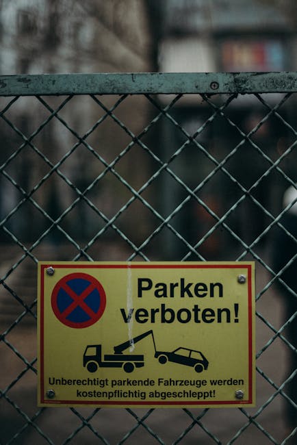 A close-up view of a metal chain-link fence with a yellow warning sign attached, located on Romford Road in Forest Gate. The sign displays a blue and red no-parking symbol and text in German, stating 'Parken verboten!' which translates to 'No parking!' and includes an illustration of a tow truck towing a car, indicating vehicles parked illegally will be towed at the owner's expense. The fence appears weathered with visible rust spots and is situated near a building in the background, with part of a window and some blurred urban elements. The setting suggests a busy street environment, often associated with house removals or relocation services, where parking restrictions are enforced to facilitate loading and moving activities. This image is relevant to local regulations affecting house removals and furniture transport in Newham, with a focus on parking restrictions specific to Forest Gate, as highlighted on the company website manandvanforestgate.co.uk.