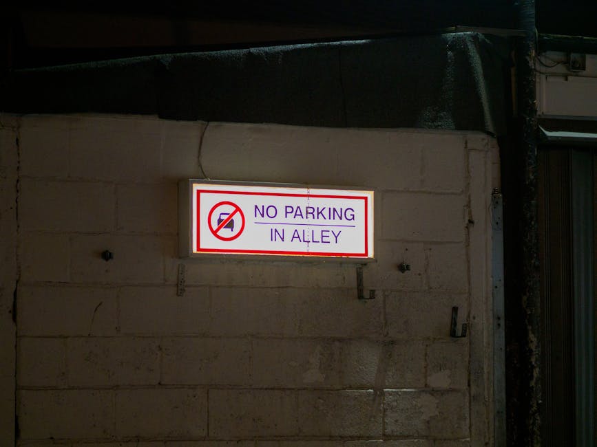 A rectangular illuminated traffic sign mounted on a beige brick wall indicates 'No Parking in Alley' with a red circle and slash over a black car symbol, situated outdoors at night. The sign features purple text and a red border, and is attached with metal brackets. To the right, part of a metal roller shutter door is visible, suggesting an industrial or commercial property. The wall appears weathered, with some cracks and uneven bricks, and ambient lighting highlights the sign's visibility. This setting could be part of a home relocation or furniture transport scenario, where understanding parking restrictions on Romford Road is essential for efficient removal services, as provided by Man and Van Forest Gate.