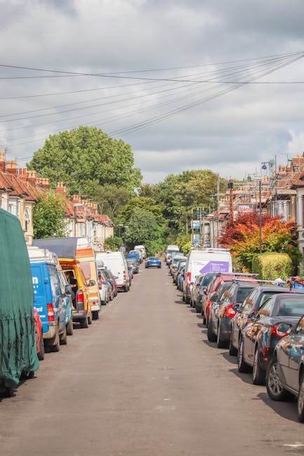 A residential street during daytime with closely parked cars on both sides, including a mix of small hatchbacks and larger vans, some of which are covered in protective wraps. The pavement is clear, suggesting a typical urban setting suitable for house removals and furniture transport. The background features rows of terraced houses with red brick facades, small front gardens, and some greenery, along with a few trees and a cloudy sky overhead. Overhead power lines run across the street, and street lamps are visible along the pavement. The scene depicts the environment in which Man and Van Forest Gate provides house removal services, including loading and unloading furniture, appliances, and boxes, with a focus on efficient movement and careful handling in tight urban spaces, aligning with the context of packing and moving logistics in Forest Gate for a home relocation.