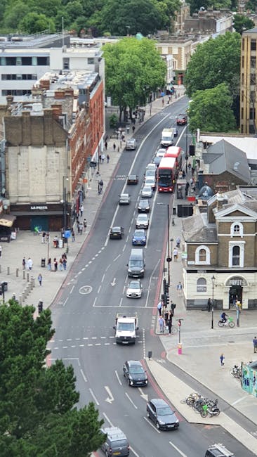 An aerial view of a busy urban street scene showing multiple vehicles including cars, vans, and a red bus moving in both directions. The street is bordered by a mix of residential and commercial buildings, with some older brick structures and modern facades. Green trees line the sidewalks, providing shade and greenery amidst the city environment. Pedestrians can be seen walking along the pavements, and bicycles are parked near a small building at the corner. The scene includes a section of pavement with a few people waiting, and a traffic intersection with marked lanes and pedestrian crossings. This setting illustrates the typical urban traffic flow and street activity that accompanies home relocation and furniture transport processes, which companies like Man and Van Forest Gate handle efficiently during removals and moving services.