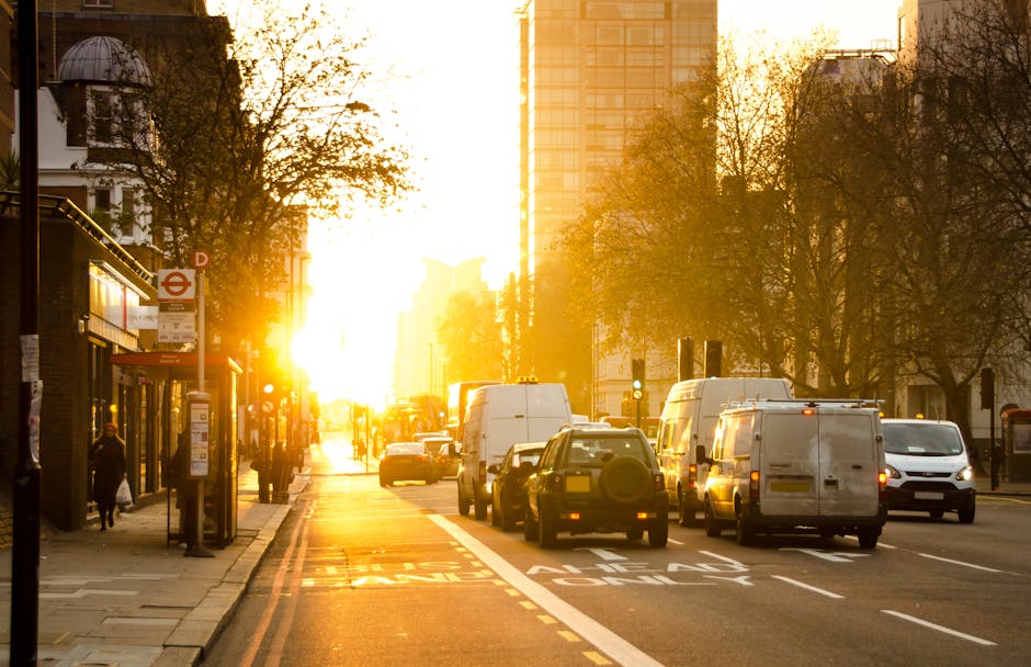 A busy urban street scene during sunset with several white and dark coloured vans, trucks, and cars parked along the roadside, some positioned for loading or unloading, indicative of home relocation and furniture transport activities. The sidewalk features a shelter with a bus stop sign and passengers waiting, while pedestrians walk past nearby shops and buildings, including a white building with large windows on the left. Tall trees line the street, casting long shadows as the golden sunlight filters through the branches, illuminating the scene. In the background, high-rise buildings reflect the sunset glow, creating a warm atmosphere typical of late afternoon or early evening. The image captures the logistical elements of a house removals operation, such as vehicle positioning and urban environment, relevant to services offered by Man and Van Forest Gate in the context of same day removals delays and solutions in Forest Gate.