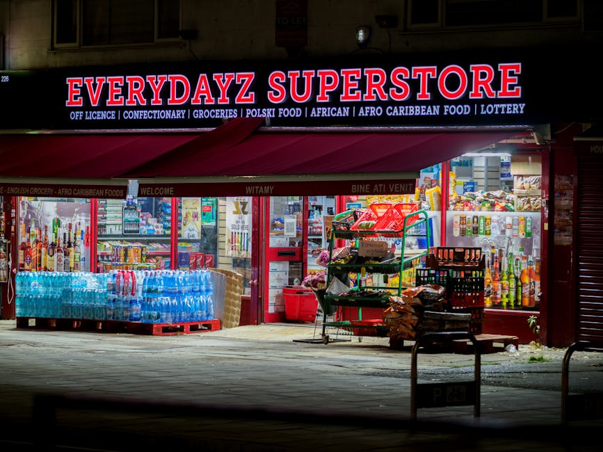 A brightly lit storefront of Everydayz Superstore at night, displaying various grocery items, bottled drinks, and snacks stacked outside on pallets and carts. In front of the shop, there are large pac