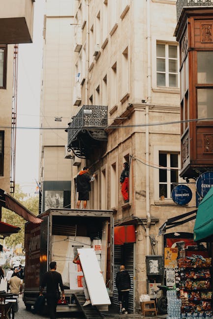 A moving scene in a narrow urban street showing the side of a multi-storey building with a small metal balcony and laundry hanging on a line. A mover is seen outside on a ladder, wearing an orange safety helmet, working on the building's exterior or installing equipment. A large moving truck is parked on the street with its rear doors open, revealing packed cardboard boxes and plastic wrapping materials inside; some boxes are stacked on the pavement nearby. Several pedestrians are visible walking or standing nearby, with some observing the activity. The surrounding area includes various storefronts with signage and awnings, and elements of street furniture and overhead power lines add to the busy city atmosphere. This scene illustrates the logistical challenges associated with tight access home relocation or furniture transport in a dense urban environment and highlights the involvement of professional removal services like Man and Van Forest Gate in residential moving projects.